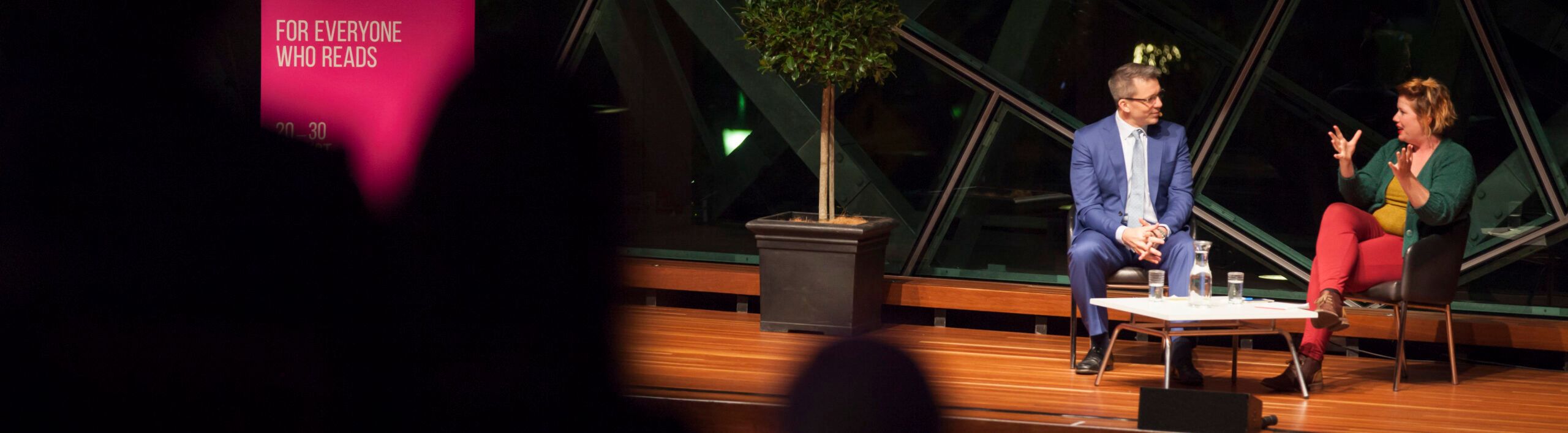 A photograph of two Melbourne Writers Festival speakers on stage, illuminated by a spotlight.