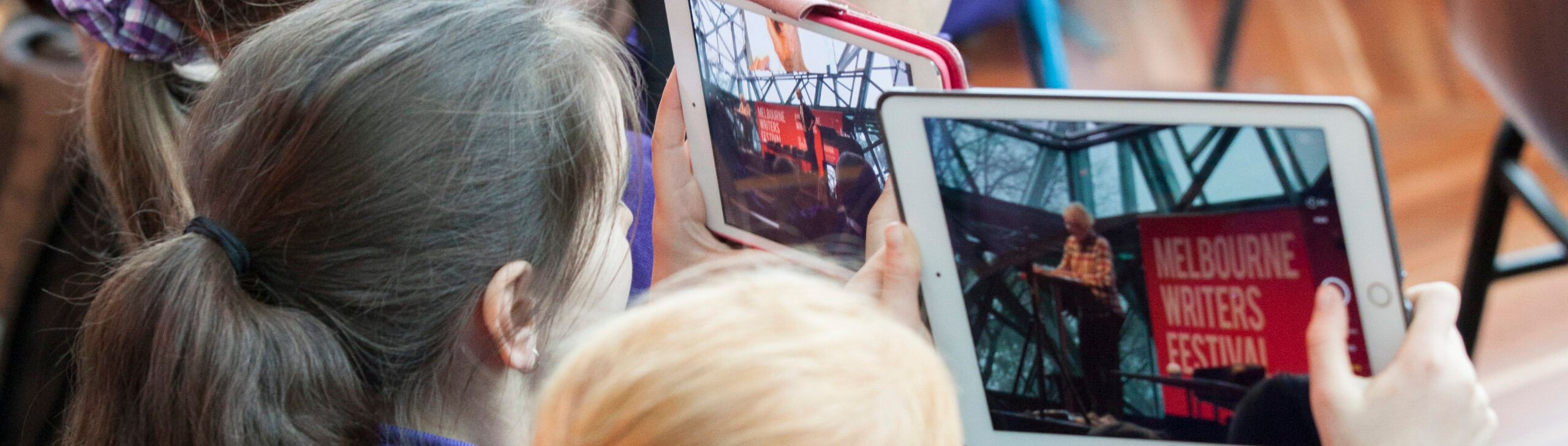 A close up of two children taking photographs of a Melbourne Writer Festival event through their iPads.